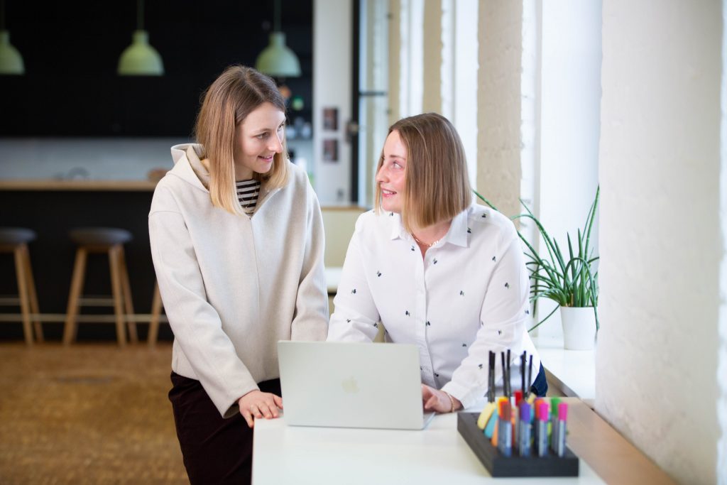 Eine Kollegin erklärt einer anderen etwas am Laptop.